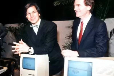 A young Steve Jobs in a bow tie with John Sculley behind two original Macintosh computers